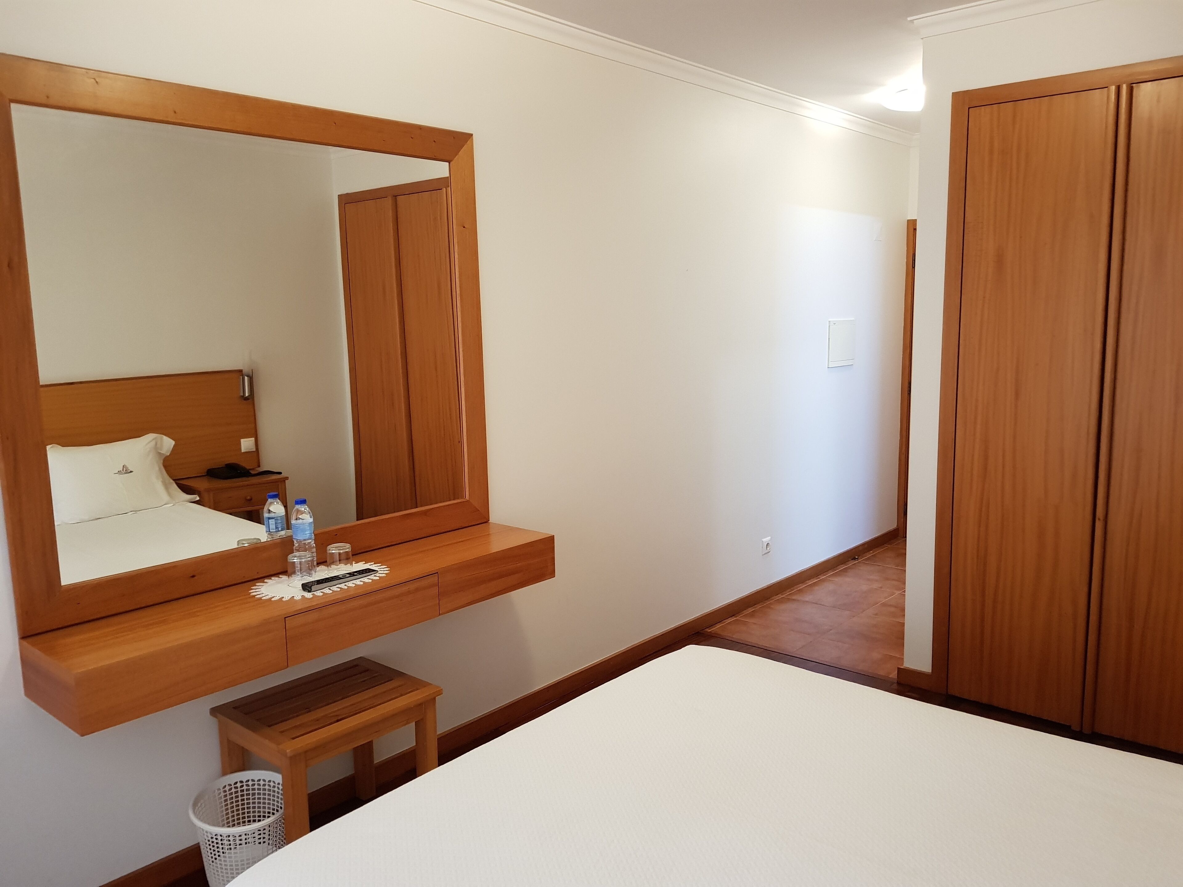 Habitación de hotel con cama de madera, una mesita de noche con teléfono y botella de agua, un taburete pequeño de madera, un cesto de ropa blanca y un espejo grande.
