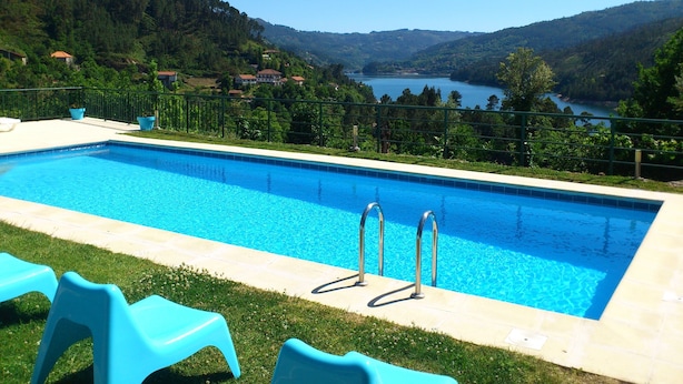 Una piscina rectangular d'aigua clara i blava, envoltada per una paret baixa de pedra. Hi ha dues cadires de plàstic blau sobre la gespa davant la piscina. La piscina fa de fons a uns turons verds i un llac.