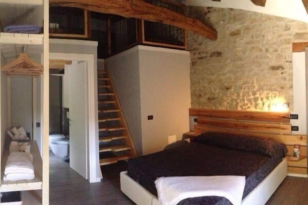 A compact bedroom with a bed, a small bathroom, a wooden staircase, and a stone wall.