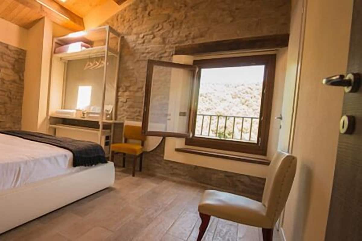 A bedroom with a stone wall, a bed, a desk, and a window with a view.
