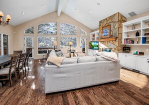 Amplia sala de estar con un sofá gris grande, una mesa de comedor de madera y sillas. Se observan ventanas amplias con vistas a árboles nevados, una chimenea de piedra y estanterías blancas con libros y objetos decorativos.