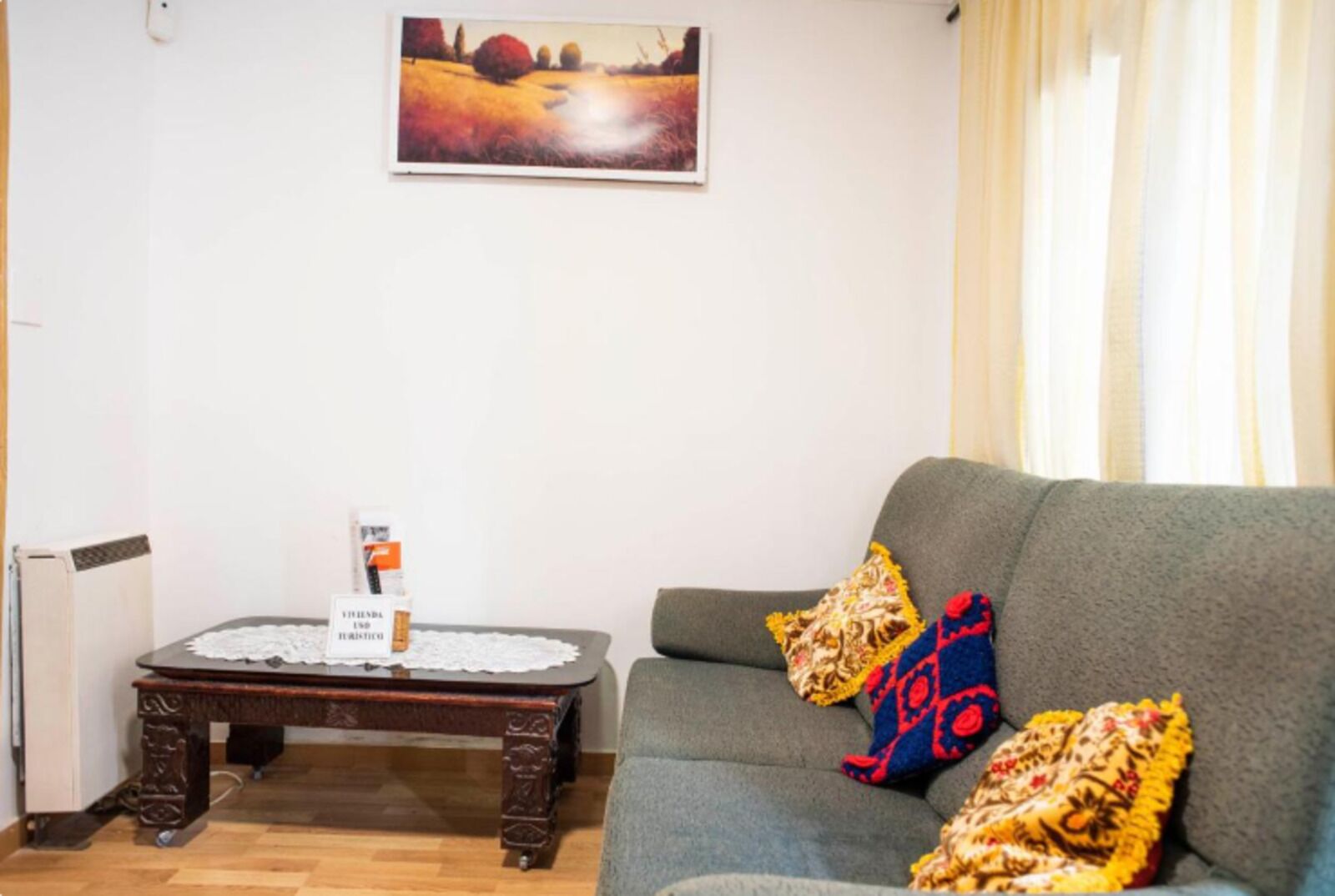 A living room with a grey sofa, two patterned throw pillows, a wooden coffee table with a white lace tablecloth, and a painting above the table.