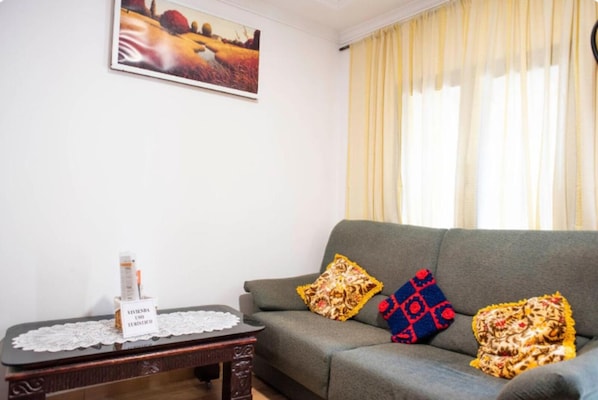 A living room with a grey sofa, two patterned cushions, a small wooden table with a lace doily, and a framed landscape painting on the wall.