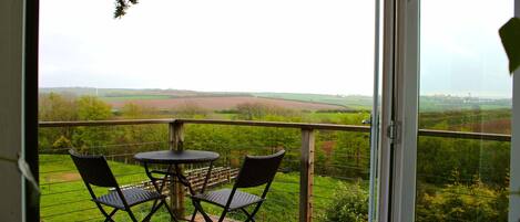 A balcony with a table and chairs, offering a view of a green landscape and fields.