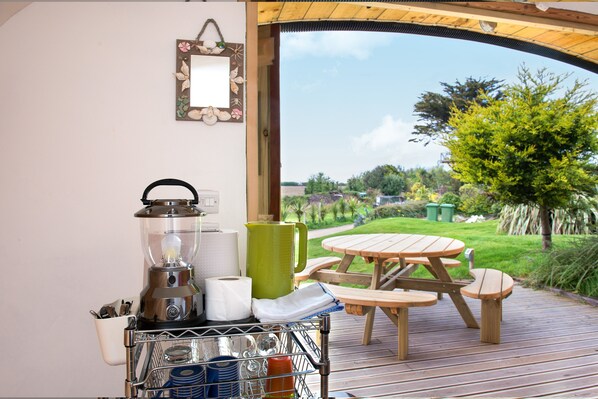 A kitchenette with a blender, kettle, and coffee maker, a wooden table and chairs, and a view of a garden. - Atlantic Surf Pods - Campsite (Bude)