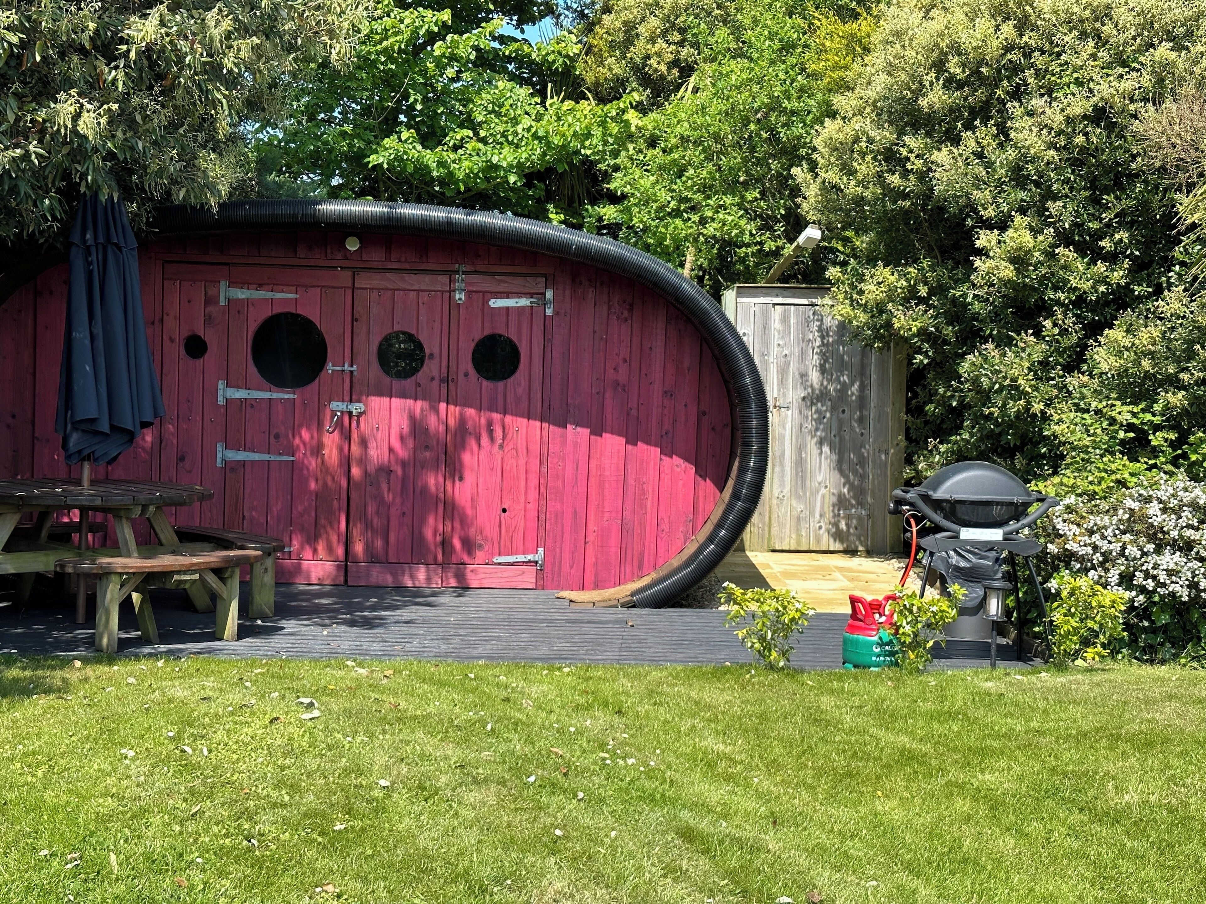Una estructura roja de madera con ventanas circulares, una mesa de picnic y una parrilla en un jardín.