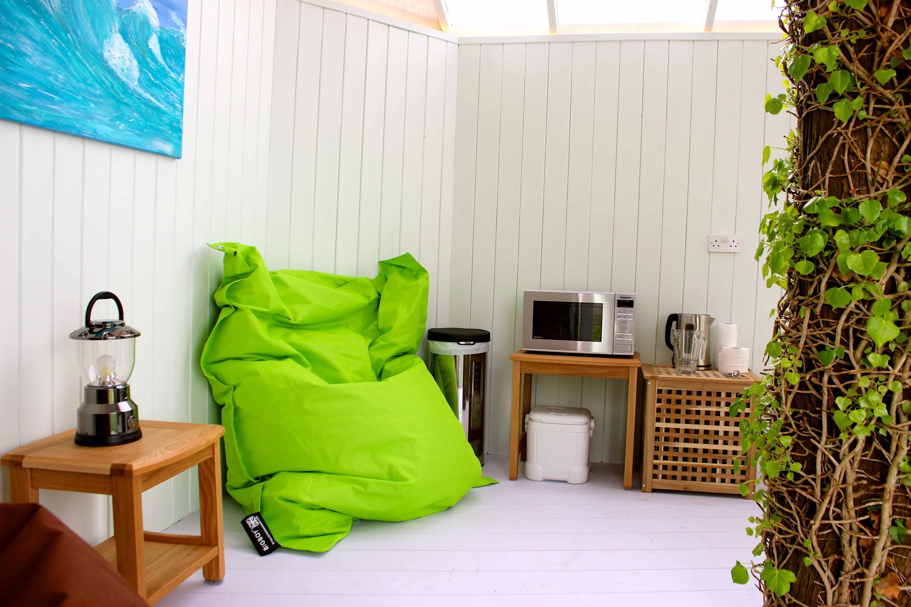 A cozy room with a green bean bag chair, a small wooden table with a lantern, a microwave, and a trash bin.