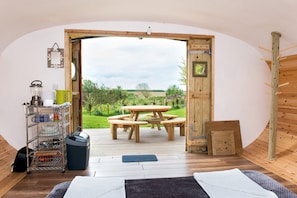 A room with a wooden dining table and benches, a metal shelf with dishes, a trash bin, and a view of a green landscape through an open door. - Atlantic Surf Pods - Campsite (Bude)