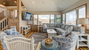 A spacious living room with a large sectional sofa, a wicker chair, and a small table with a basket. There is a staircase with wooden railings leading to an upper level. - Dancing Dolphin | 240 ft from the beach | Private Pool, Hot Tub, Dog Friendly | Duck (Duck)