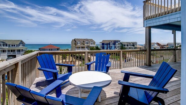 A wooden deck with blue Adirondack chairs and a white table, overlooking a beach with houses and the ocean. - Dancing Dolphin | 240 ft from the beach | Private Pool, Hot Tub, Dog Friendly | Duck (Duck)
