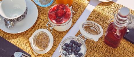 A breakfast setup with a woven placemat, a cup of coffee, a glass of water, a jar of yogurt with berries, a glass of juice, a bottle of syrup, and a plate with croissants.