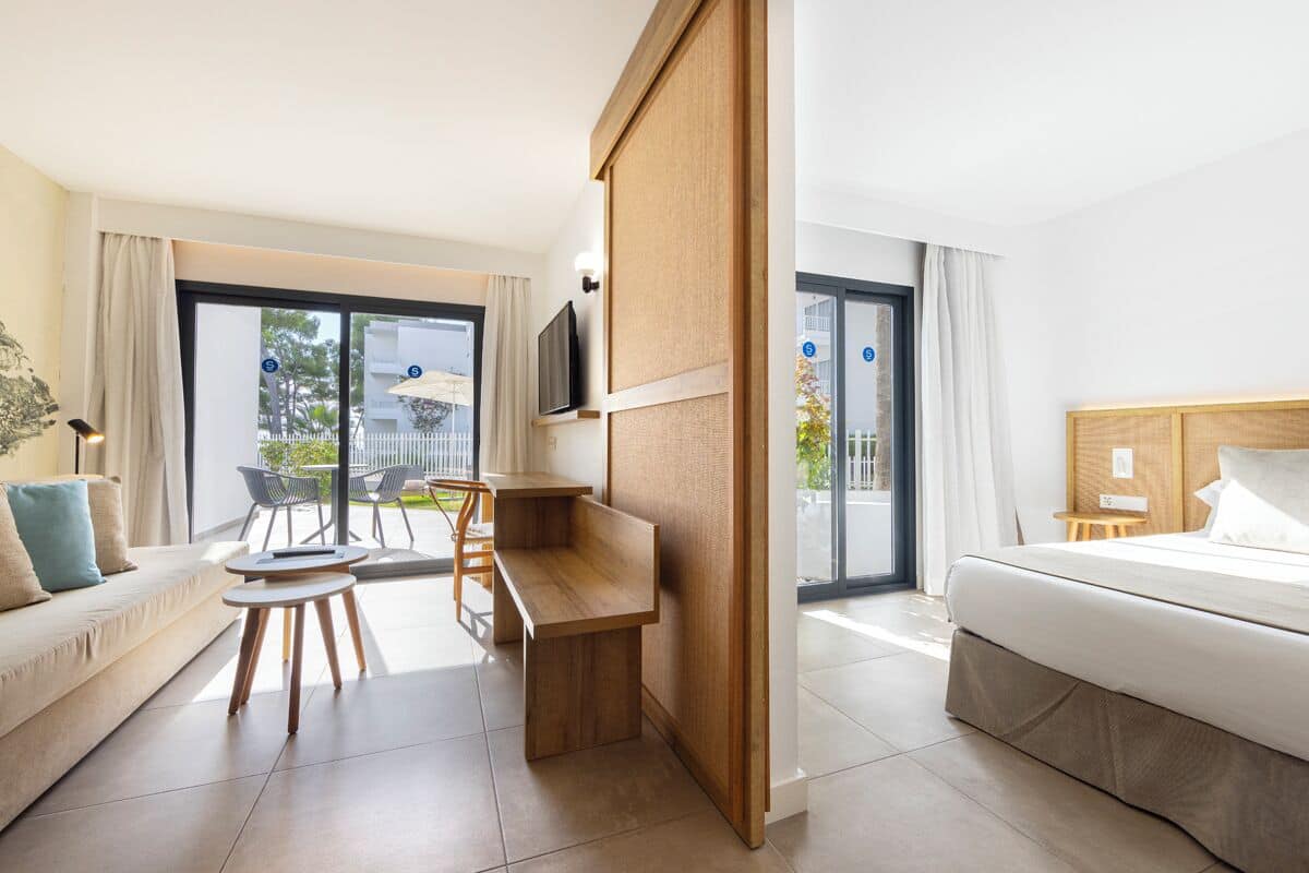 Quarto de hotel moderno com cama, sofá, escrivaninha e varanda com mobiliário exterior.