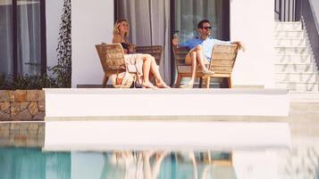 Una pareja descansando junto a una piscina con sillones de mimbre, un muro de piedra y un edificio moderno al fondo.