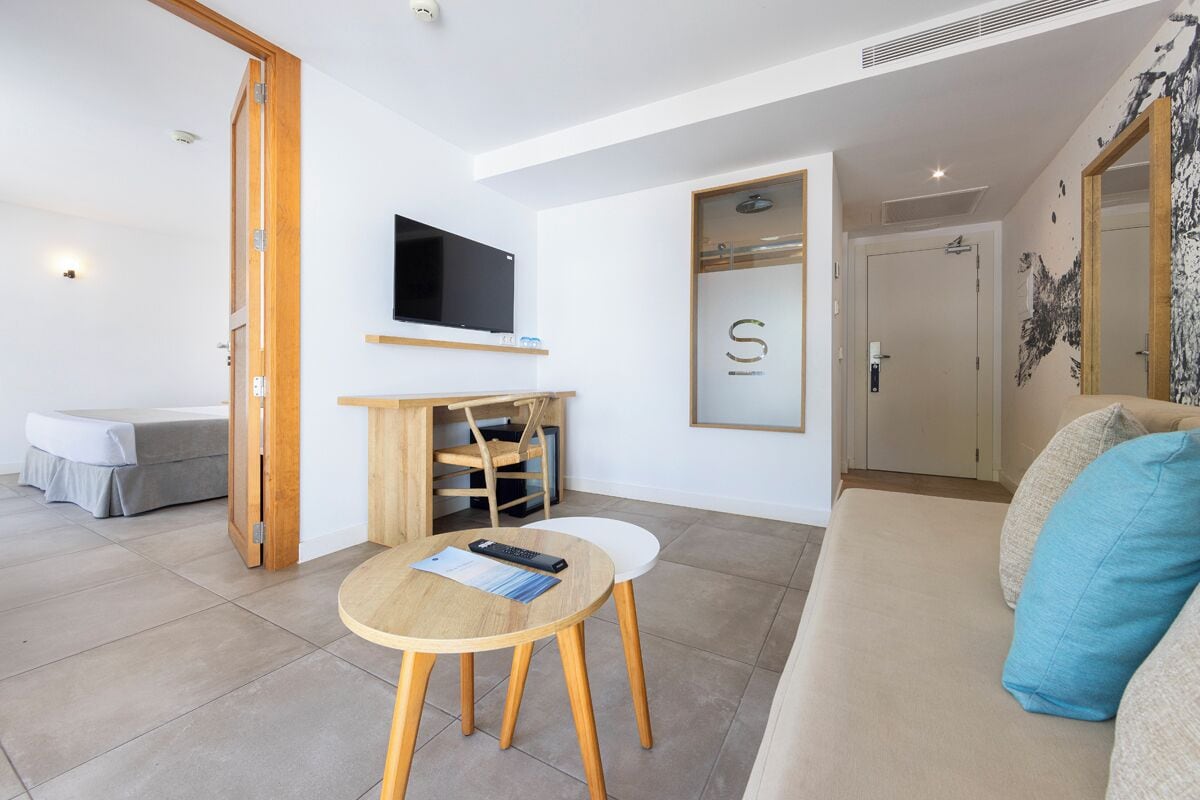 Uma sala de estar moderna com sofá, mesa redonda de madeira, TV fixada na parede e uma porta com uma placa com a letra 'S'.