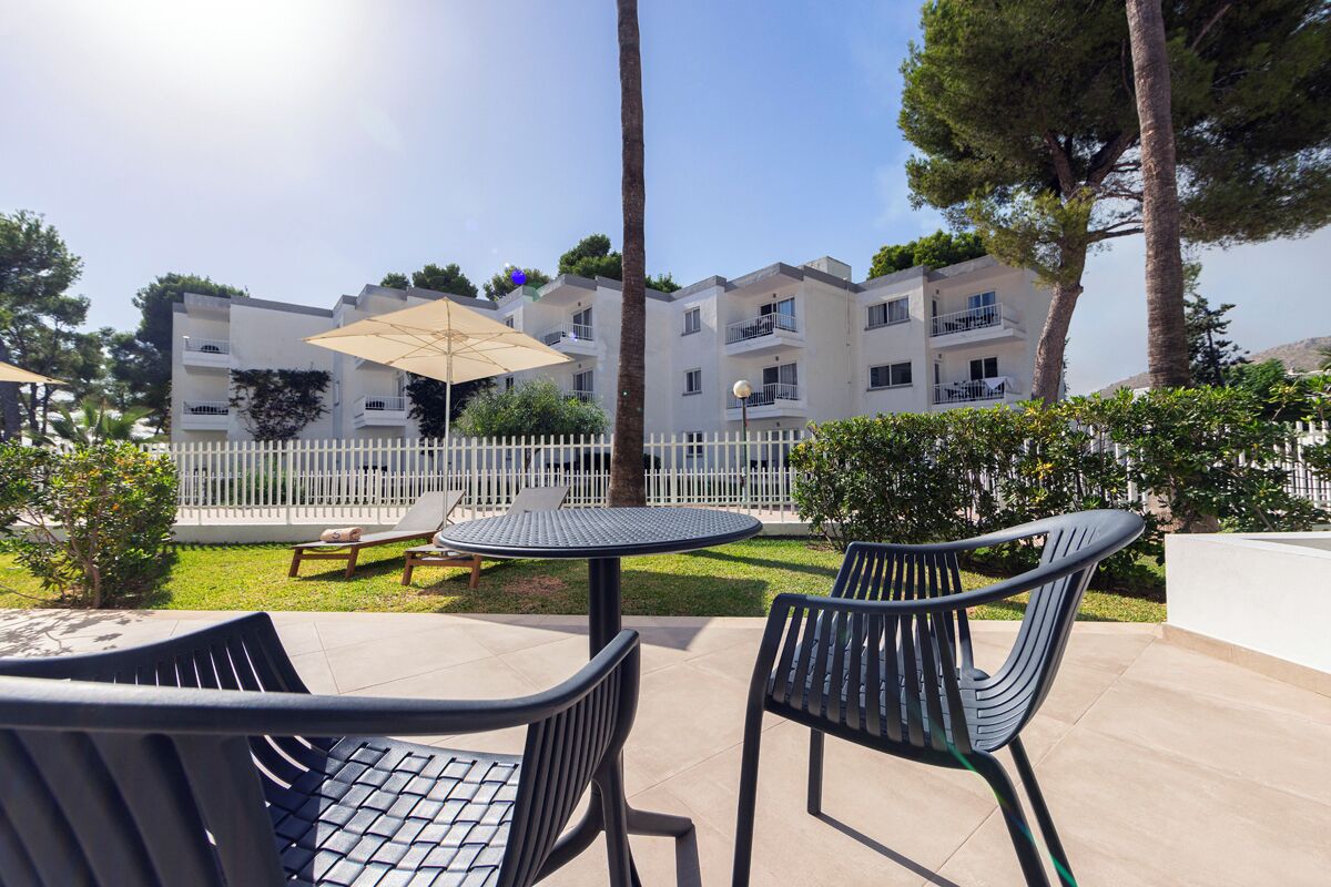 An outdoor patio with a round table and two chairs, a white fence, and a building with balconies in the background.