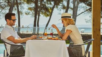 Dos personas cenando en una mesa al aire libre junto a una piscina, con vista al mar y árboles.