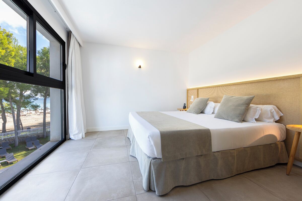 Een moderne hotelkamer met een groot bed, een nachtkastje en uitzicht op het strand door middel van ramen van vloer tot plafond.