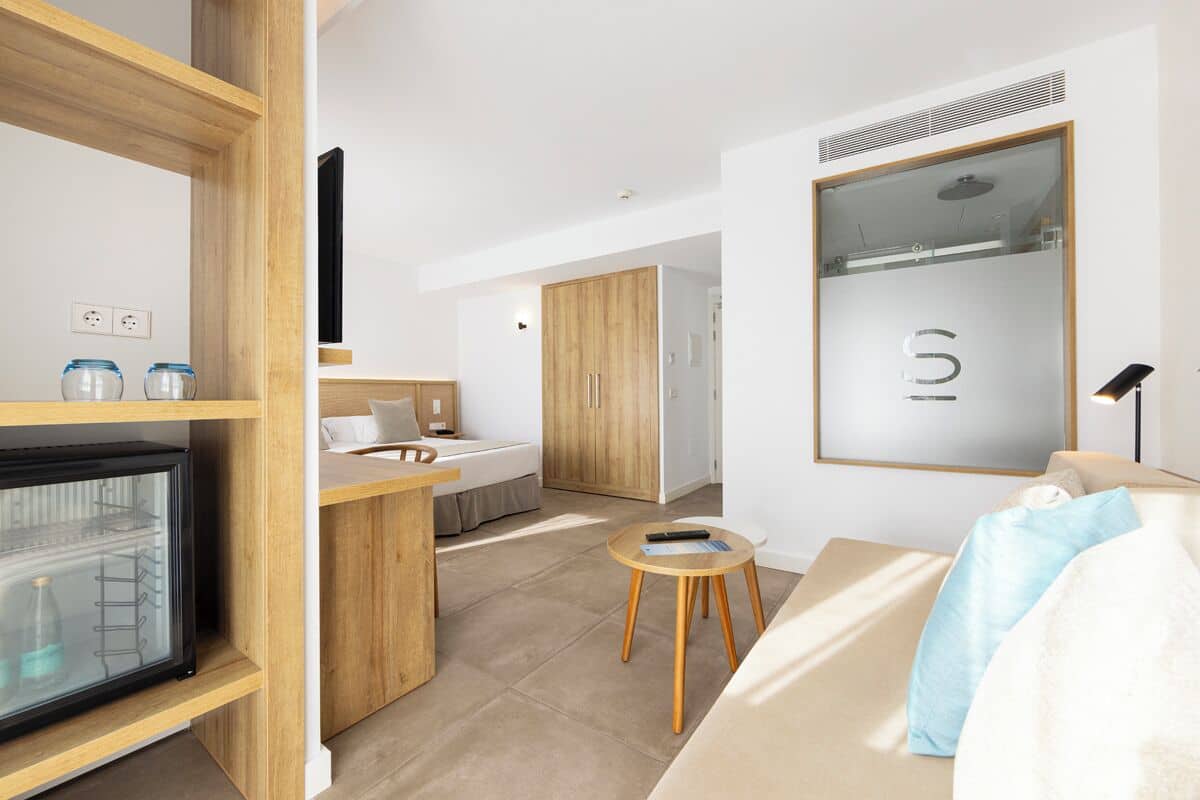 Quarto de hotel moderno com cozinha compacta, cama com travesseiro branco e azul, mesa redonda pequena e um espelho grande com o logo 'S'.