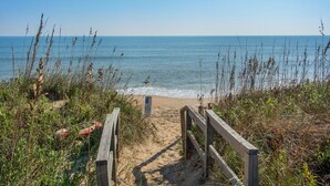 Ein Holzsteg führt zu einem Strand mit klarem, blauem Wasser und sandigem Ufer.