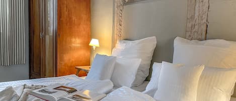 A neatly made bed with white linens, a wooden wardrobe, a bedside table with a lamp, and a bowl of fruit on the bed.