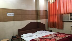 Un dormitorio con cabecera de madera, cama con ropa de cama blanca y una colcha floral roja, mesita de noche con un teléfono, un taburete pequeño y un aire acondicionado instalado en la pared.