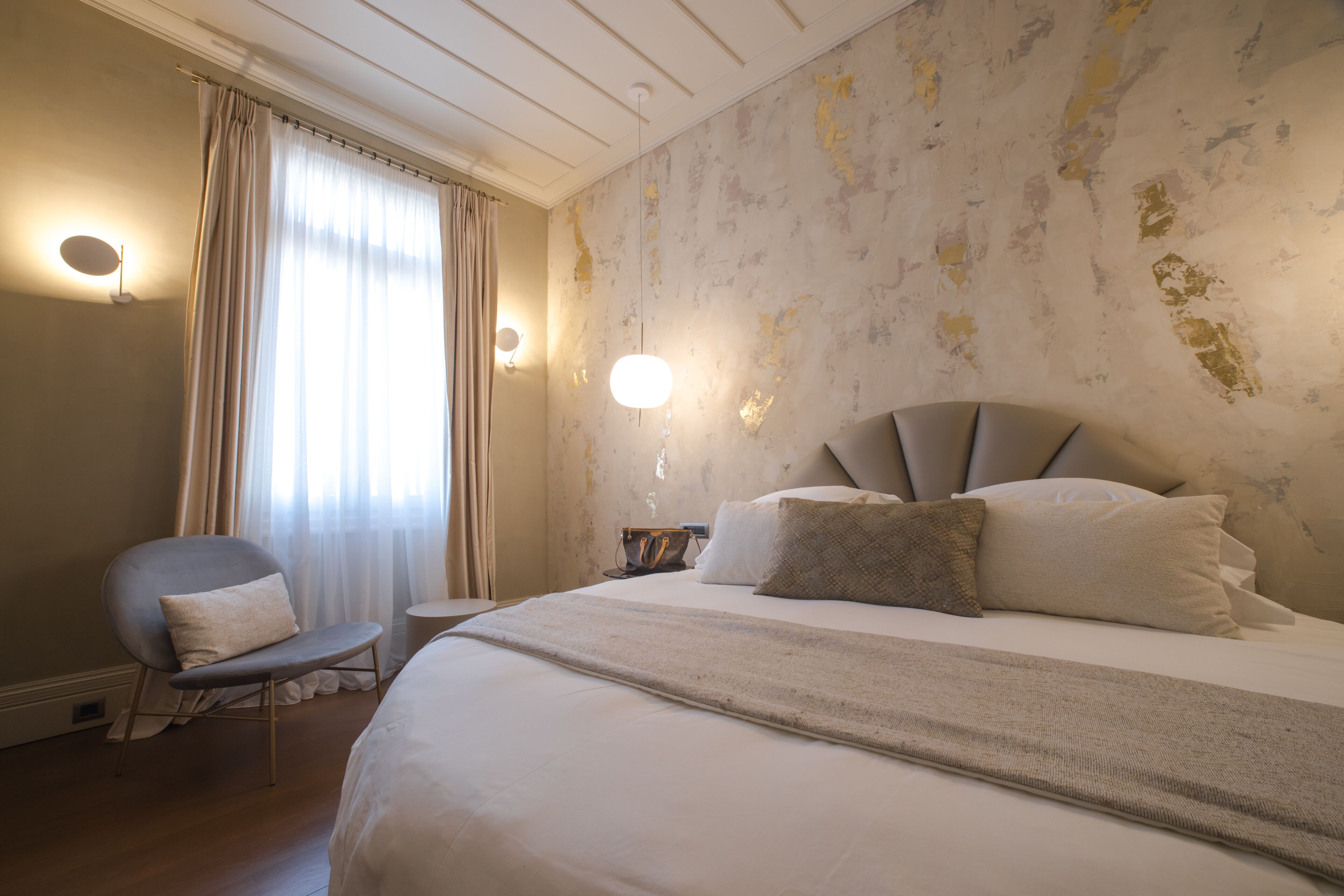Habitación de hotel con una cama grande, una silla, una mesita de noche y una lámpara de pared.