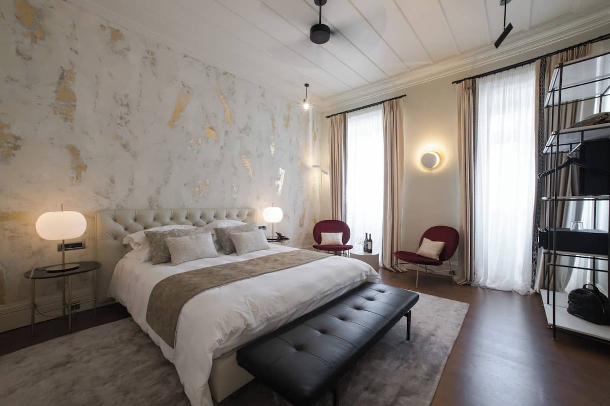 A bedroom with a large bed, bedside lamps, a bench, a red chair, a bedside table, and a wardrobe.