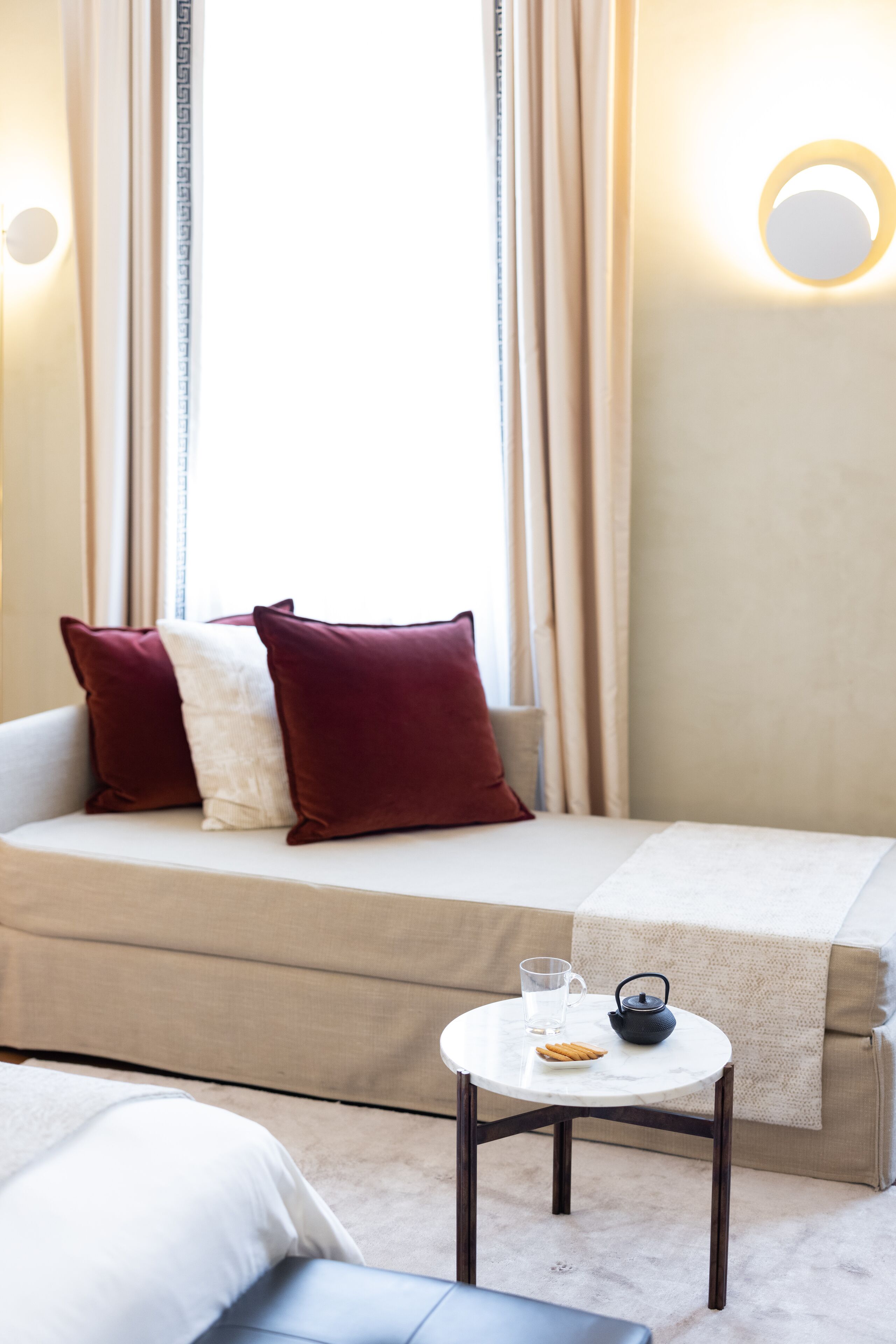 Quarto de hotel com sofá-cama, uma mesinha redonda com um bule e um copo, e janela com cortinas.