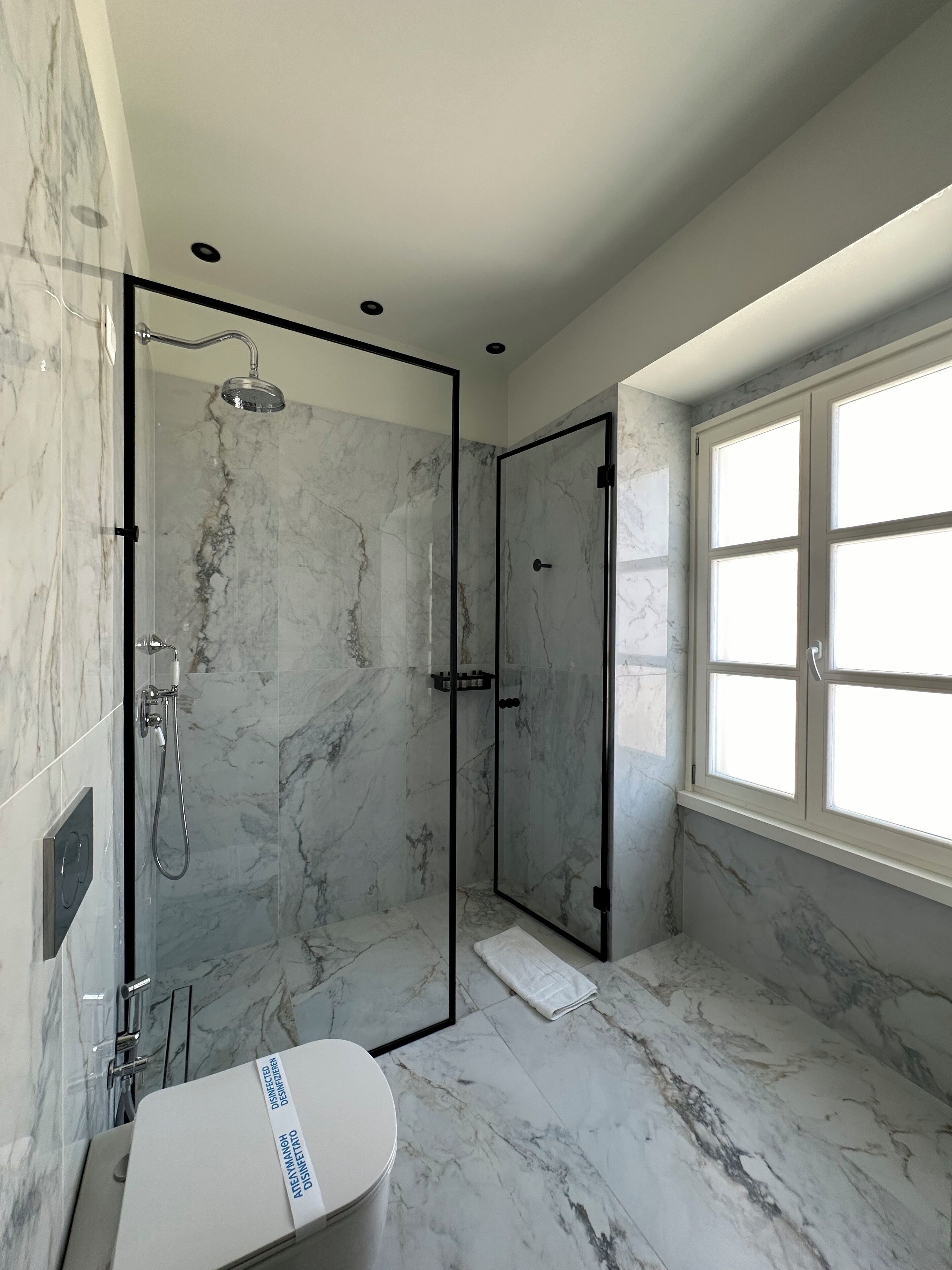 A modern bathroom with a glass-enclosed shower, marble flooring, and a large window providing natural light.
