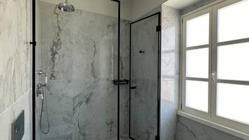 A modern bathroom with a glass-enclosed shower, marble flooring, and a large window providing natural light.