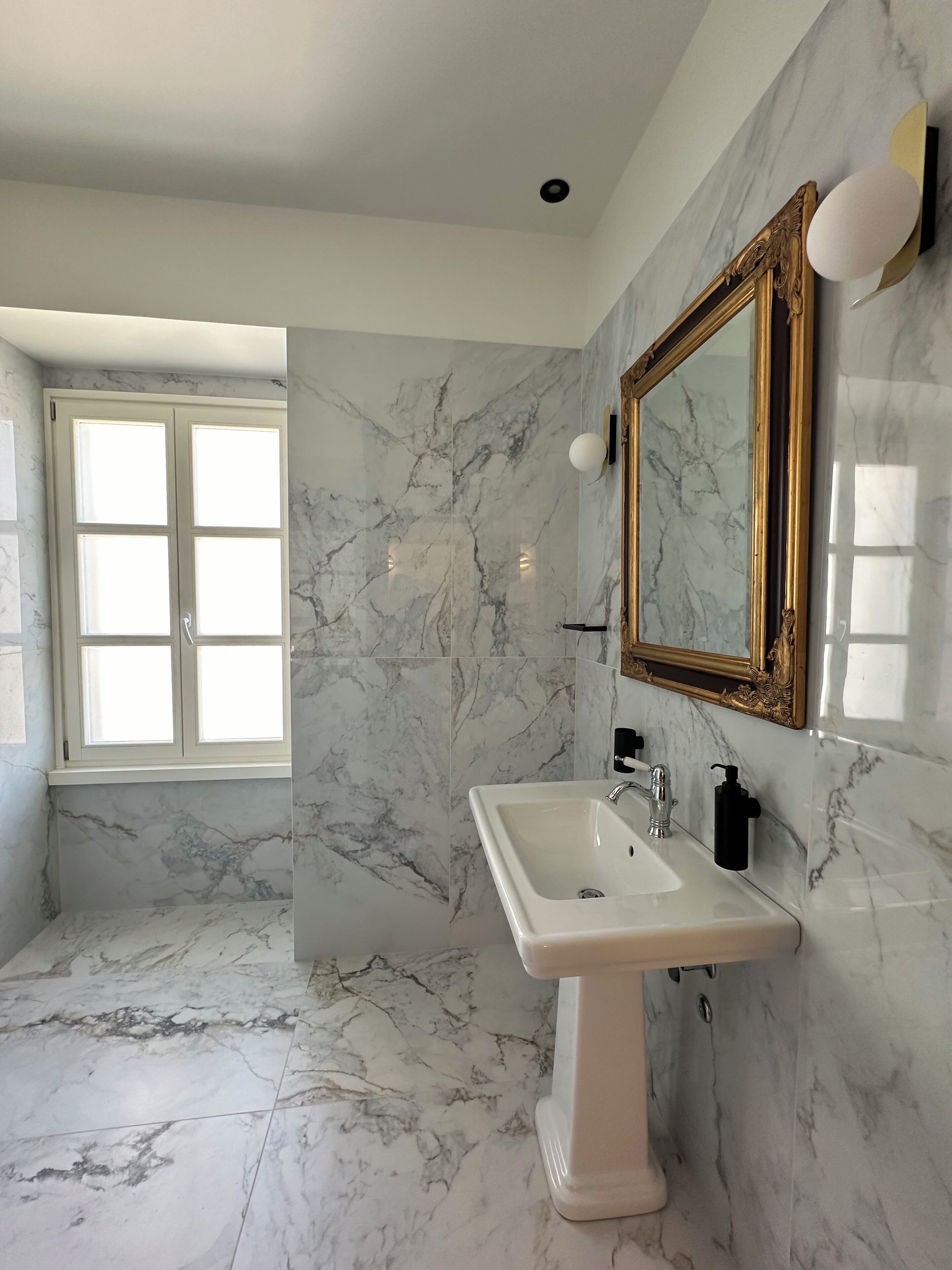 Une salle de bain aux murs et au sol recouverts de marbre, un lavabo sur pied, un grand miroir avec un cadre doré et une fenêtre avec des volets blancs.