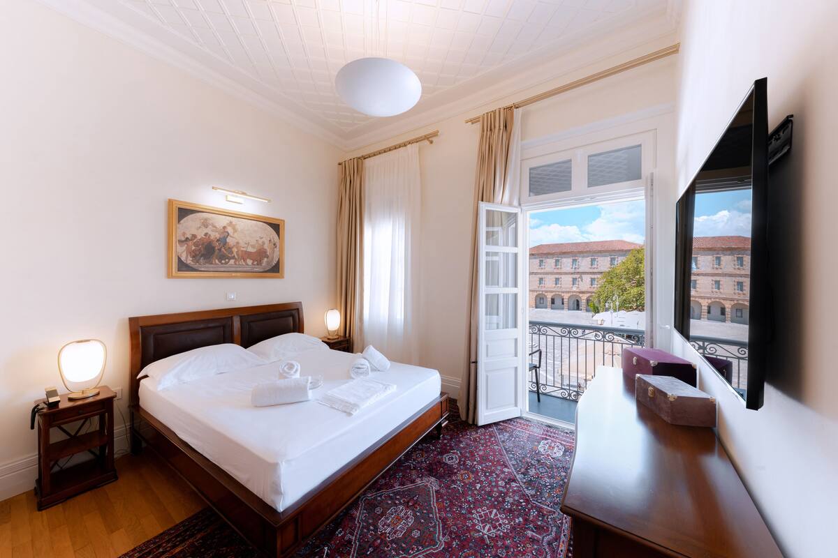 A hotel room with a bed, bedside lamps, a painting, a balcony with a view of buildings, and a desk with a chair.