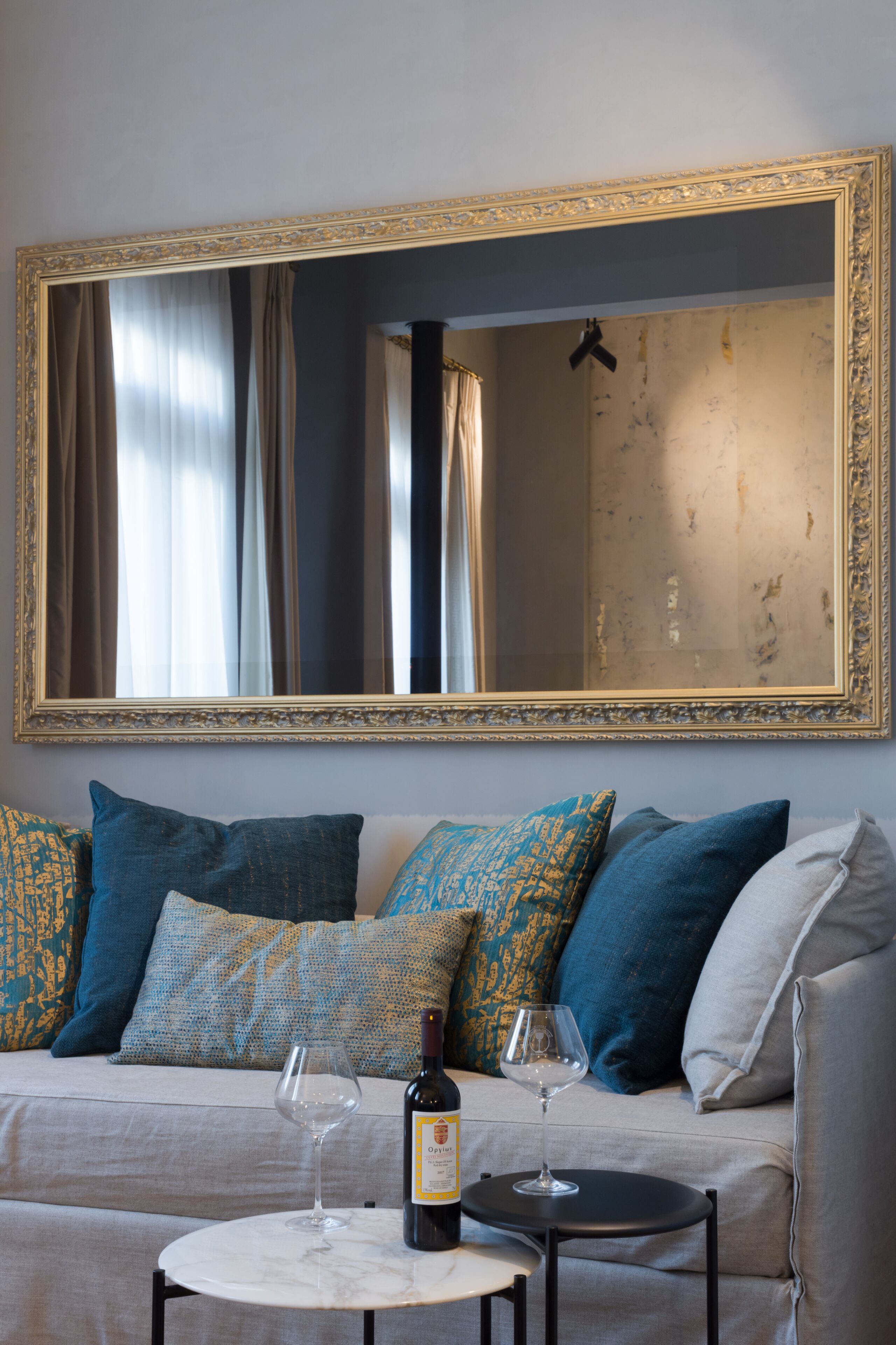 A living room with a large mirror, a sofa with patterned cushions, and two wine glasses on a small table.