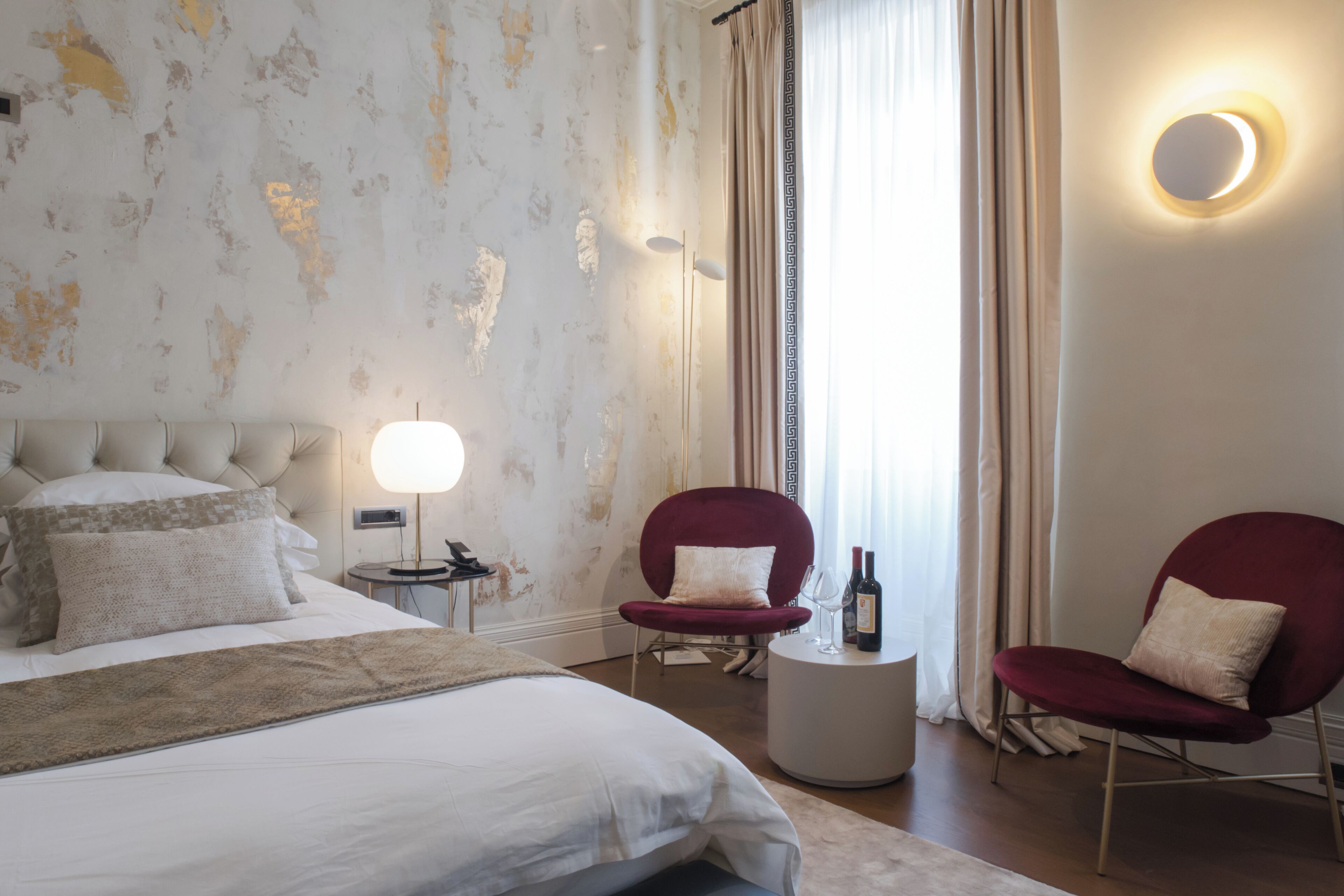 Habitación de hotel con una cama grande, dos sillones rojos, una mesita redonda con una botella y vasos, una lámpara y un ventanal con cortinas.