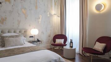 Habitación de hotel con una cama grande, dos sillones rojos, una mesita redonda con una botella y vasos, una lámpara y un ventanal con cortinas.