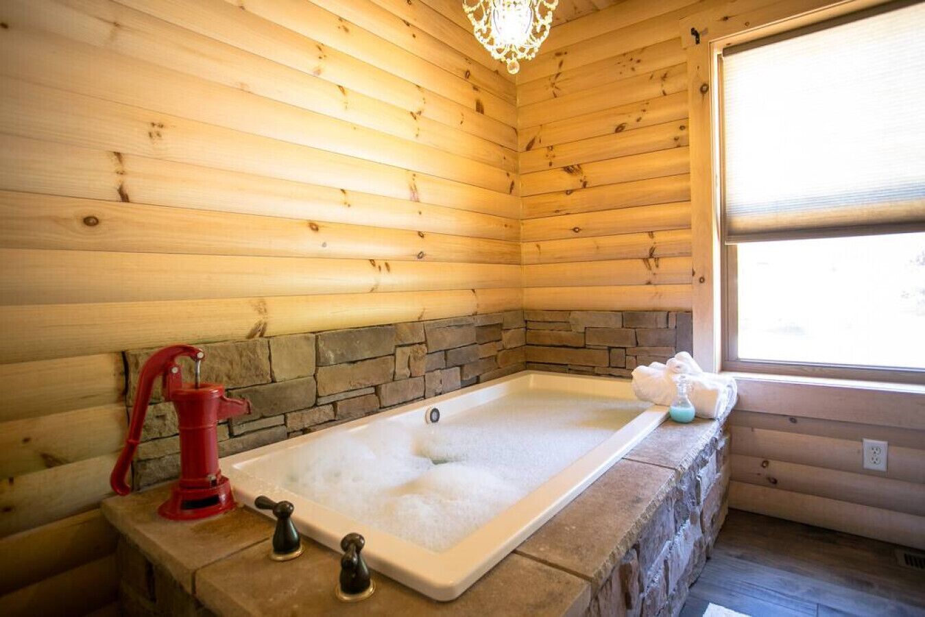 A wooden cabin interior with a stone-lined bathtub, a red faucet, and a window.