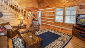 A log cabin interior with a staircase, leather furniture, a television, and a large window. - Rustic Cabin in Heart of Boone (Boone)