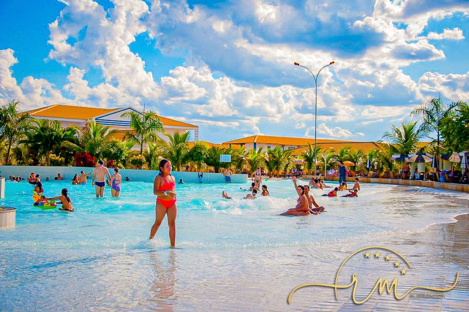 A swimming pool area with people swimming and playing, surrounded by palm trees and buildings with orange roofs.