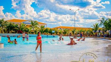 A swimming pool area with people swimming and playing, surrounded by palm trees and buildings with orange roofs.