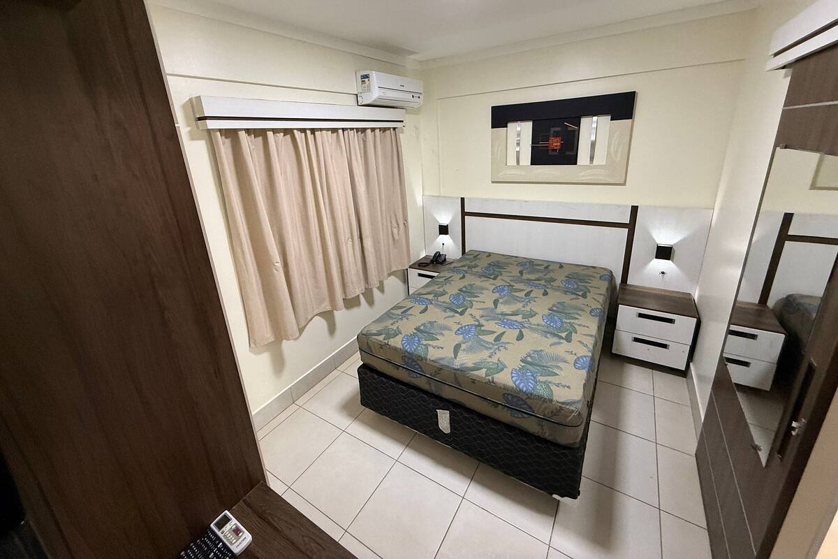 A bedroom with a bed, bedside tables, an air conditioning unit, a mirror, and a curtain.