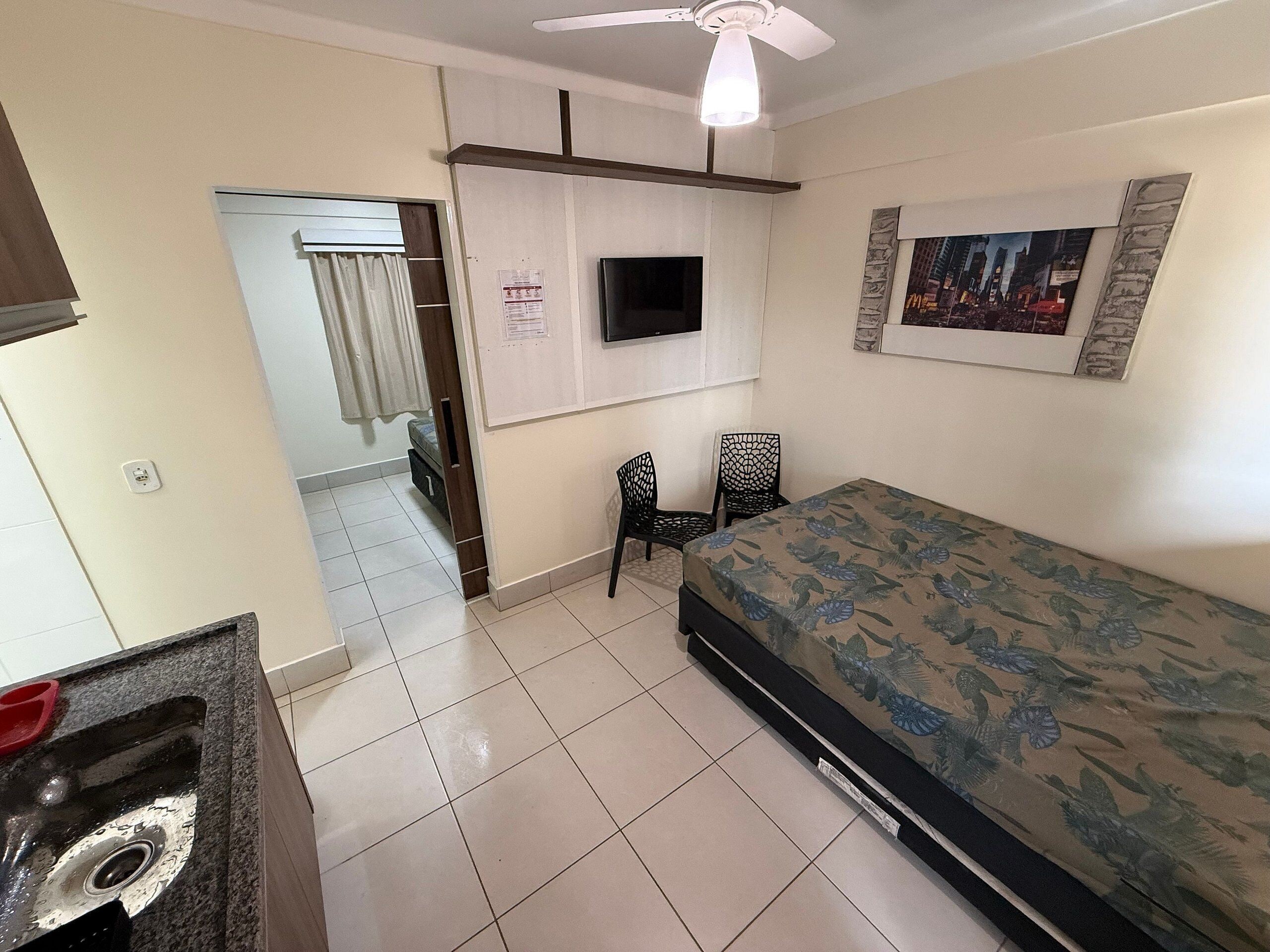 Un dormitorio con una cama, un televisor montado en la pared, un ventilador de techo y una puerta que da a otra habitación.