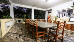 A dining area with a long table and wooden chairs, surrounded by a tiled floor and a balcony with plants. - Chácara Bosque dos Pássaros - Santa Isabel (Santa Isabel)