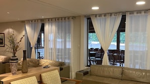 Un salon comprenant un canapé, deux fauteuils, une table basse et une table à manger avec des chaises. De grandes fenêtres sont présentes, ornées de rideaux légers, et offrent une vue sur un espace extérieur aménagé pour se détendre.