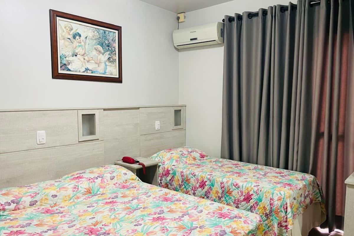 Habitación de hotel con dos camas, cubrecamas estampados con motivos florales, un cuadro en la pared, un equipo de aire acondicionado y cortinas grises.