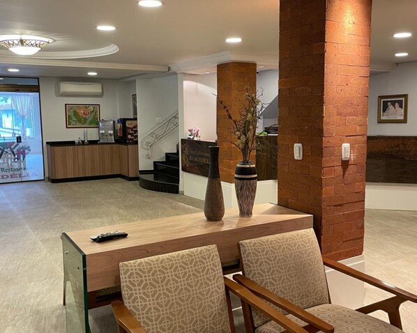 Un hall d'hôtel avec un comptoir d'accueil en bois, deux fauteuils à motifs, un vase contenant des fleurs séchées et un escalier en arrière-plan.