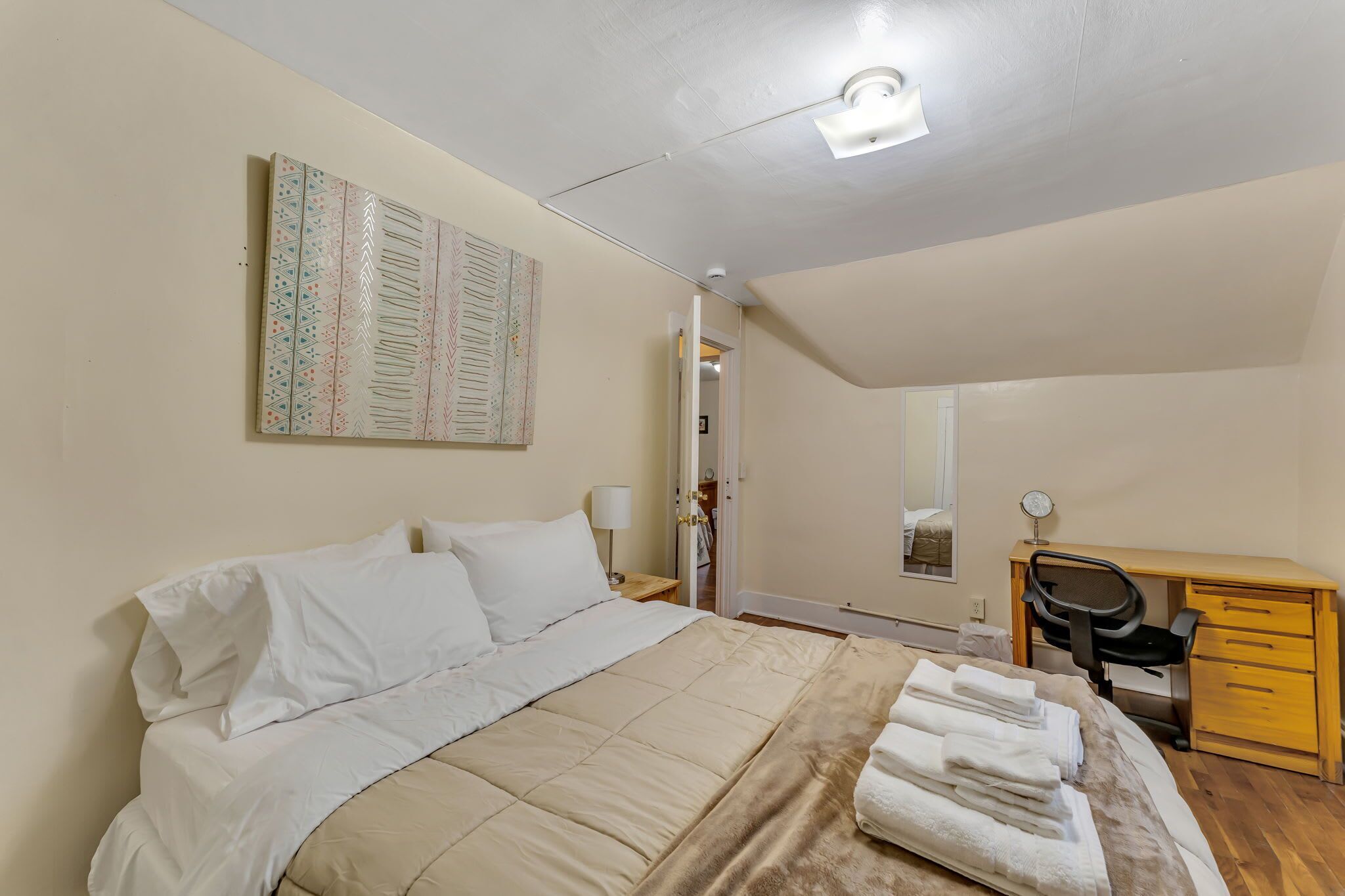A bedroom with a bed, a desk, a chair, a lamp, and a wall-mounted artwork.