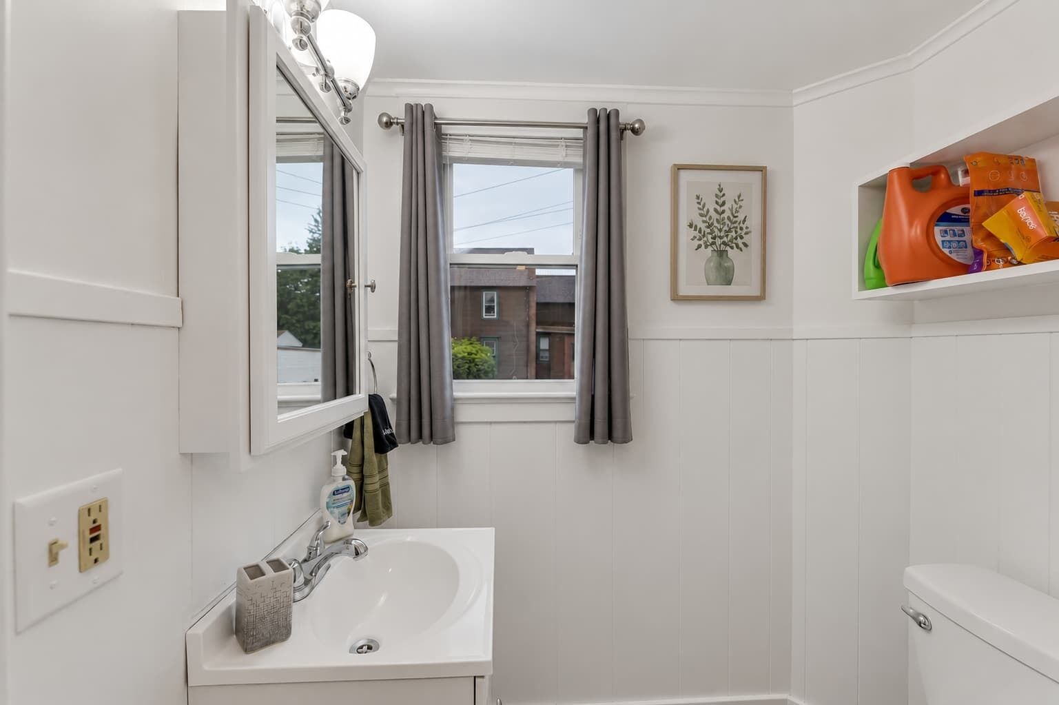 A bathroom with a white sink, a mirror, a window with grey curtains, a framed picture on the wall, and a shelf with toiletries.