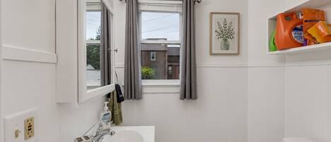 A bathroom with a white sink, a mirror, a window with grey curtains, a framed picture on the wall, and a shelf with toiletries.