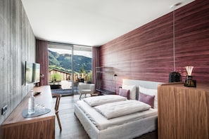 A modern hotel room with a large bed, a desk with a chair, a TV, and a view of the mountains. - Senses Violett Suites - Adults only (Zell am See)