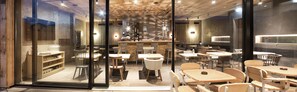 A modern cafe with wooden tables and chairs, a bar area, and a large window. - Senses Violett Suites - Adults only (Zell am See)
